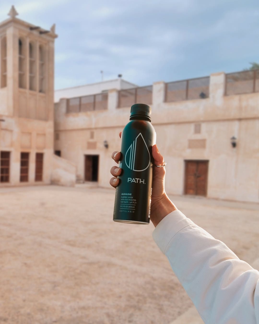 A hand holding a black PATH bottle in a sunlit Bahraini courtyard